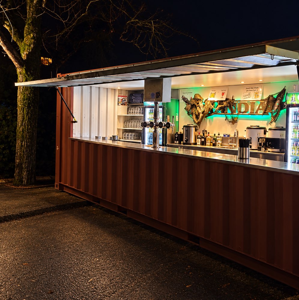 Beleuchtete Bar im umgebauten Schiffscontainer bei Nacht, mit Zapfhahn, Gläserregal und Kühlschränken