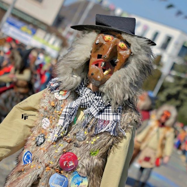 Nahaufnahme einer geschnitzten Holzmaske mit schwarzem Hut und Plakettenweste