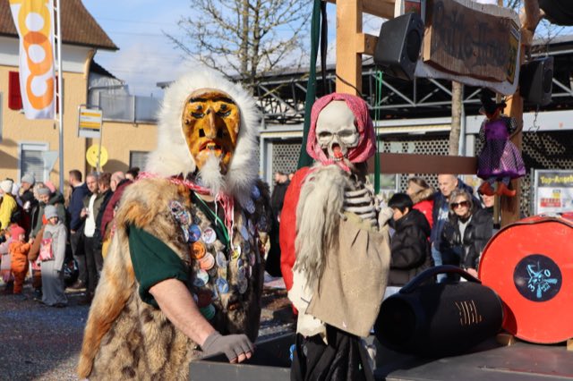 Häx mit Holzmaske steht neben einem Skelett am Wagen der Bütte Häxe