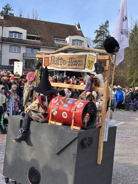 Wagen der Bütte Häxe mit Holzschild und rotem Fass, dahinter das Publikum
