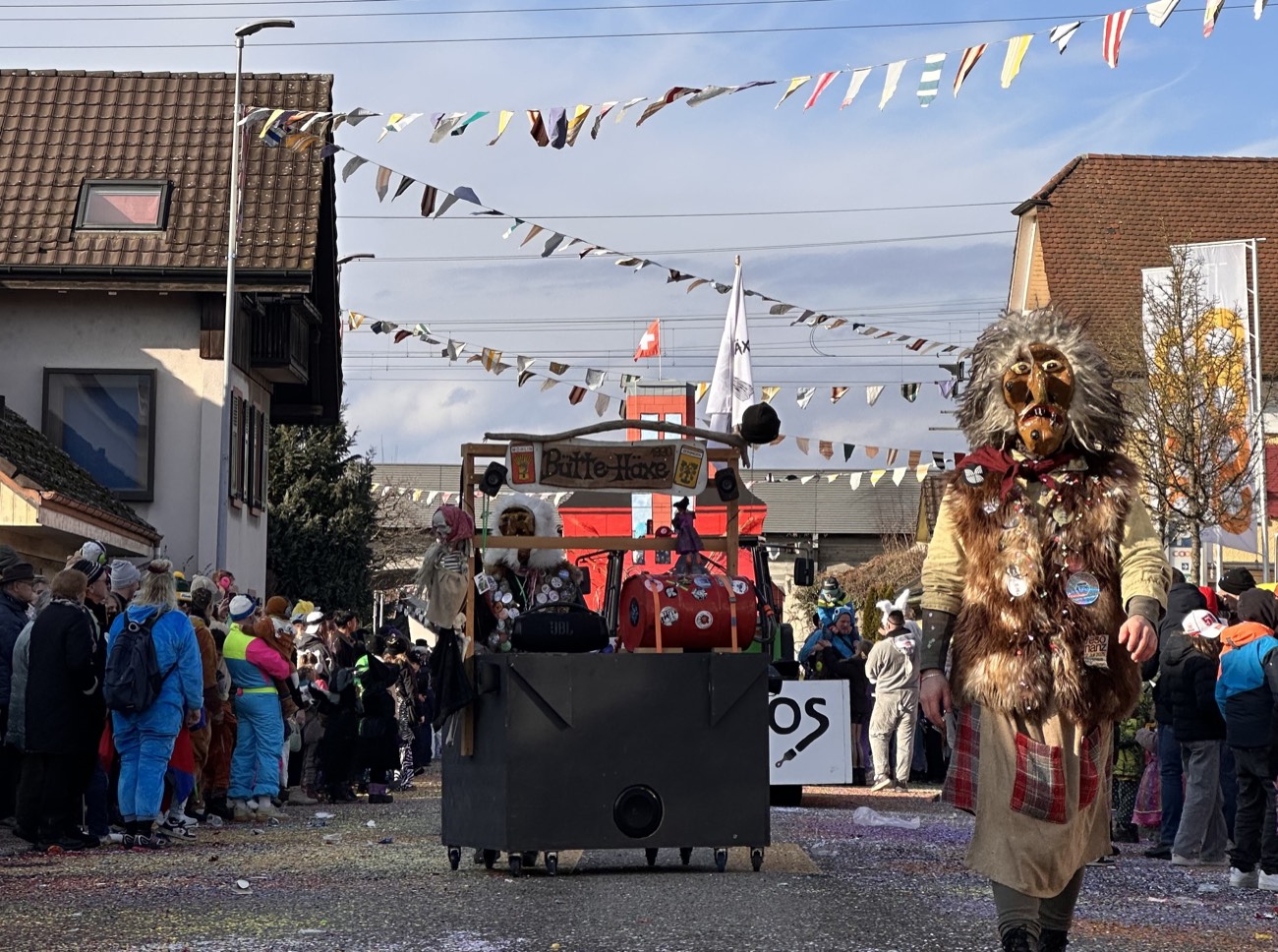 Häx mit Fellmaske vor dem Wagen der Bütte Häxe mit dem Schriftzug Bütte-Häxe, darüber bunte Wimpelketten über der Strasse in Möhlin